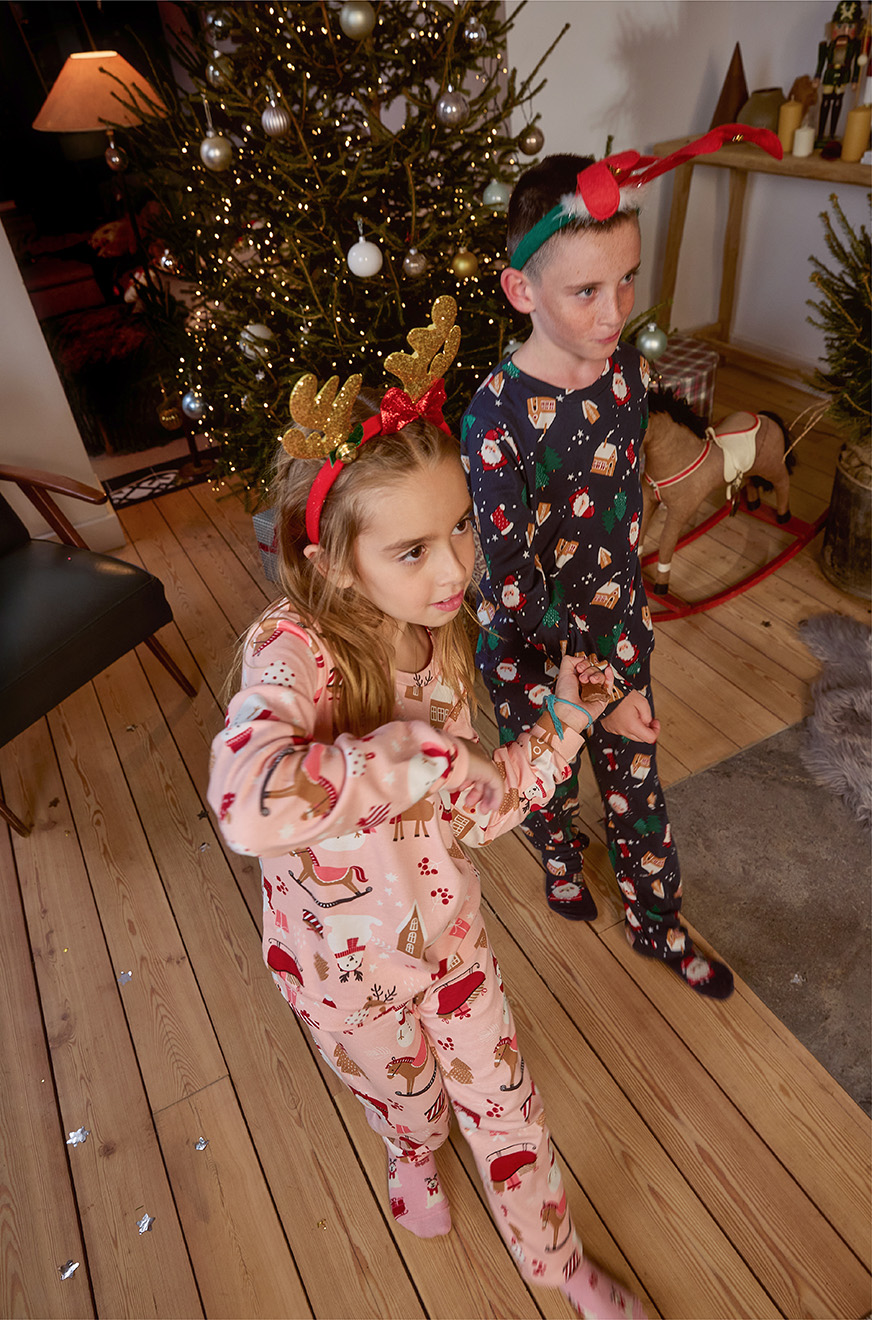 Djevojčica i dječak stoje pored jelke u pidžamama sa novogodišnjim motivima