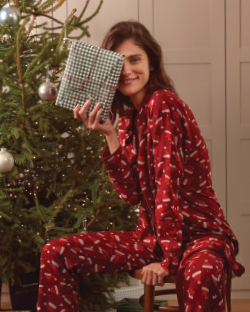 Žena u novogodišnjoj pidžamami sedi pored jelke i drži poklon